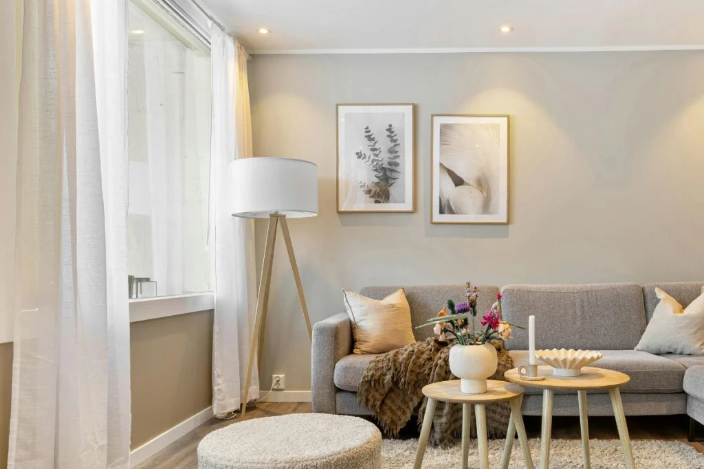 Cozy neutral living room with a gray sofa, sheer curtains, a tripod floor lamp, framed botanical prints, and round wooden side tables
